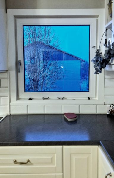 Köksfönster med vy av ett hus utanför i blått ljus. En liten låda på bänken.