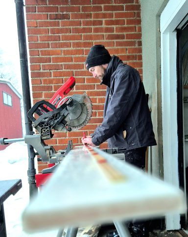Man arbetar med en såg på en balkong, med snöigt landskap i bakgrunden.